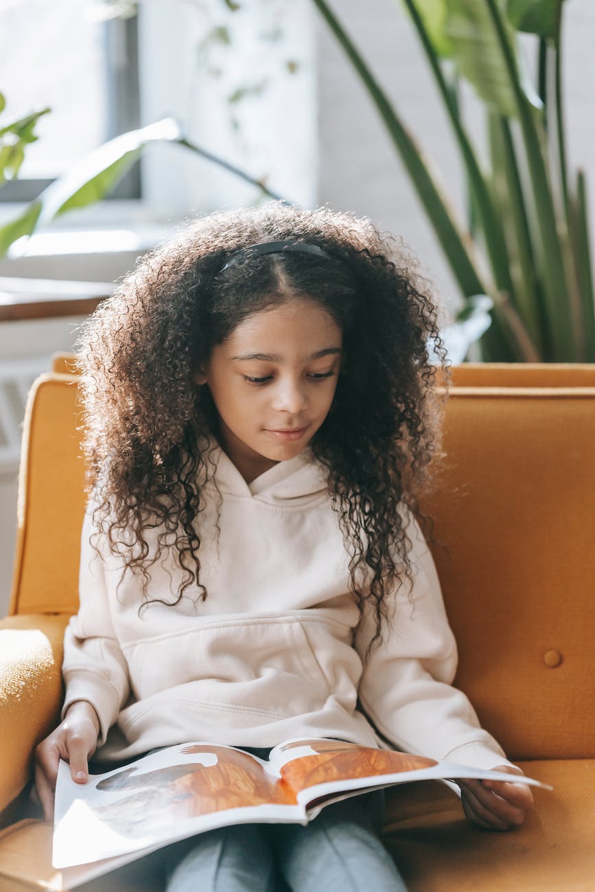 calm little ethnic girl turning book page on sofa